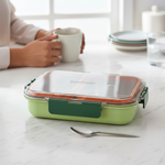 Green stainless steel food container with clear lid on kitchen table, person holding mug nearby