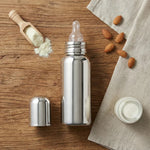 Load image into Gallery viewer, Stainless steel baby bottle with nipple and cap on wooden surface, surrounded by baby milk powder, almonds, and cloth