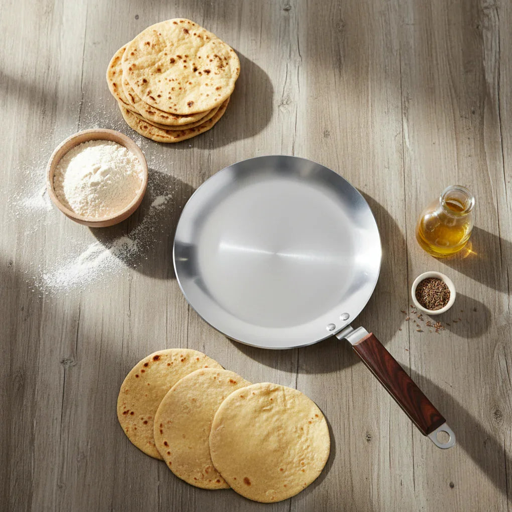 Stainless steel tawa with wooden handle on wooden table surrounded by chapati, flour, oil, and cumin seeds
